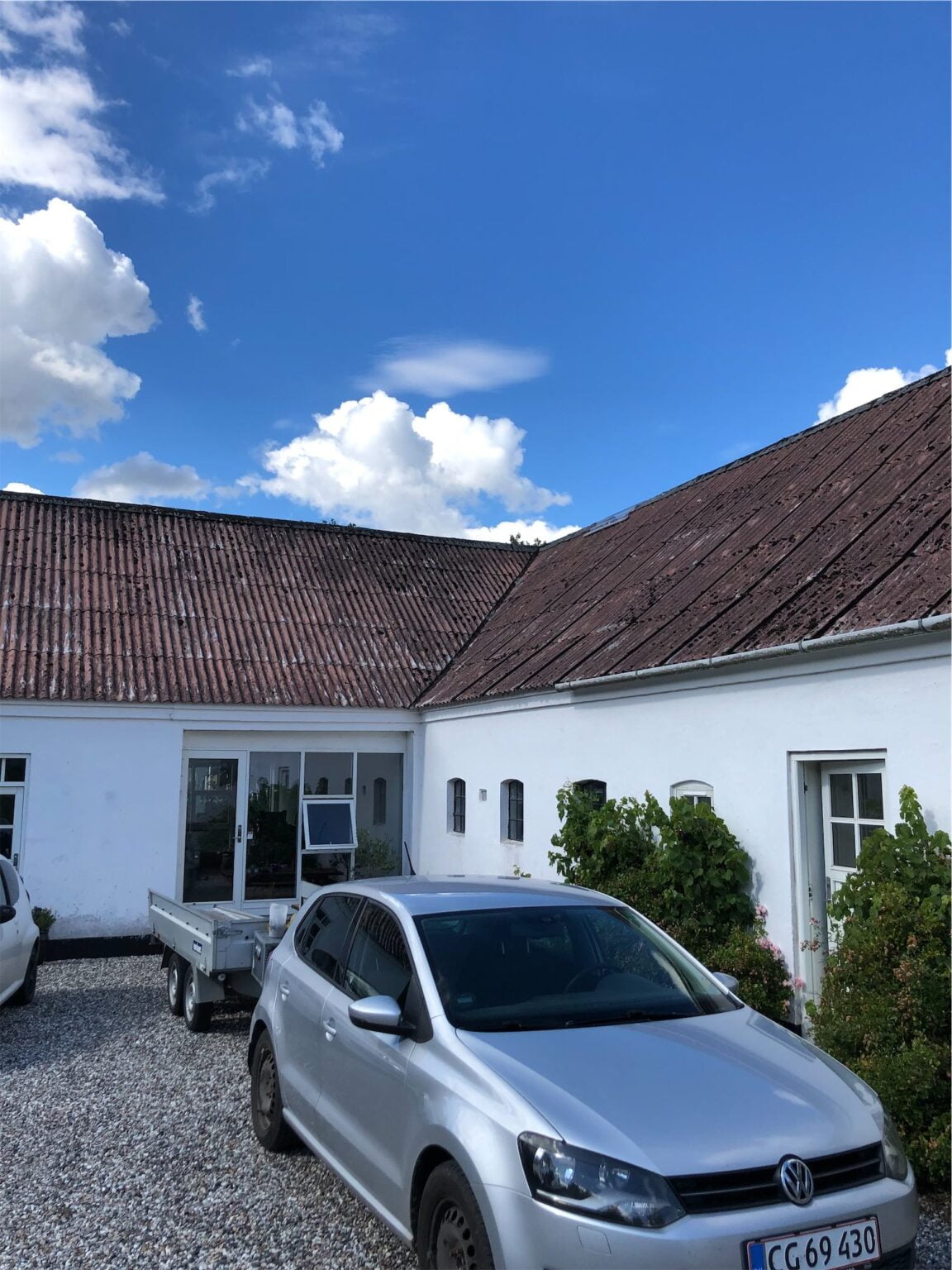 Sølv bil foran hvid bygning med blåt himmel.